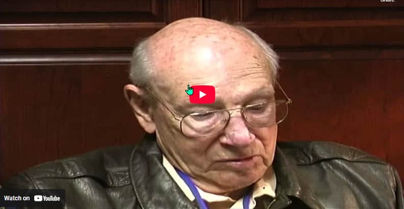 The image depicts an elderly individual with thinning gray hair, wearing glasses and a dark leather jacket. The person appears to be seated in front of a wooden paneled background, possibly in a formal setting such as a hearing or meeting. A "Watch on YouTube" button is overlaid on the image, indicating that it is a still from a YouTube video.